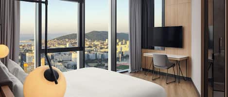 A modern hotel room with a large bed, a desk, a chair, a TV, and a view of a cityscape through large windows.