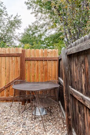 A metal table and chairs set up against a wooden fence in a garden area with gravel ground.