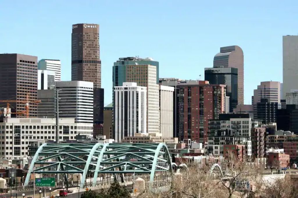 Une vue sur les toits d’une ville, avec des immeubles modernes de grande hauteur, dont un qui se distingue par un grand panneau « Qwest ».