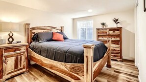 A wooden bed with a dark mattress and pillows, flanked by wooden nightstands with lamps, in a room with wooden flooring and a window with blinds. - Sloans Lake Getaway Near Broncos & Downtown Denver (Lakewood)