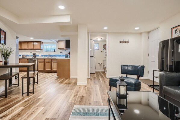 A modern kitchen with wooden cabinets, a dining area with a table and benches, and a living room with a leather chair and a TV.