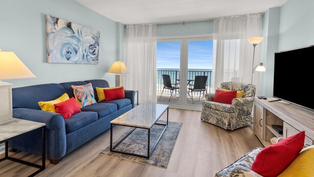 Un soggiorno moderno con un divano blu, una poltrona fantasia e una televisione. È presente un balcone con vista sull'oceano.