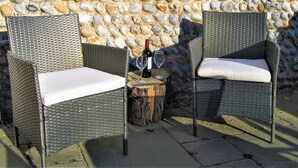 Two wicker chairs with white cushions, a bottle of wine, and a glass on a wooden stump, set against a stone wall. - 3 bedroom accommodation in Knapton, near North Walsham (Knapton, near North Walsham)