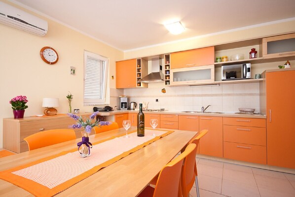 Een kleine keuken met oranje kastjes, een houten eettafel en een klok aan de muur.