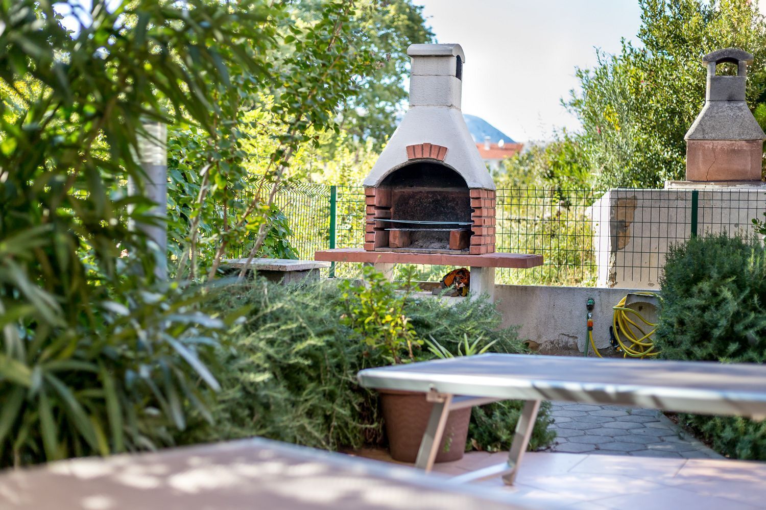 Forno de tijolos ao ar livre com chaminé, rodeado por vegetação e uma mesa de metal em primeiro plano.