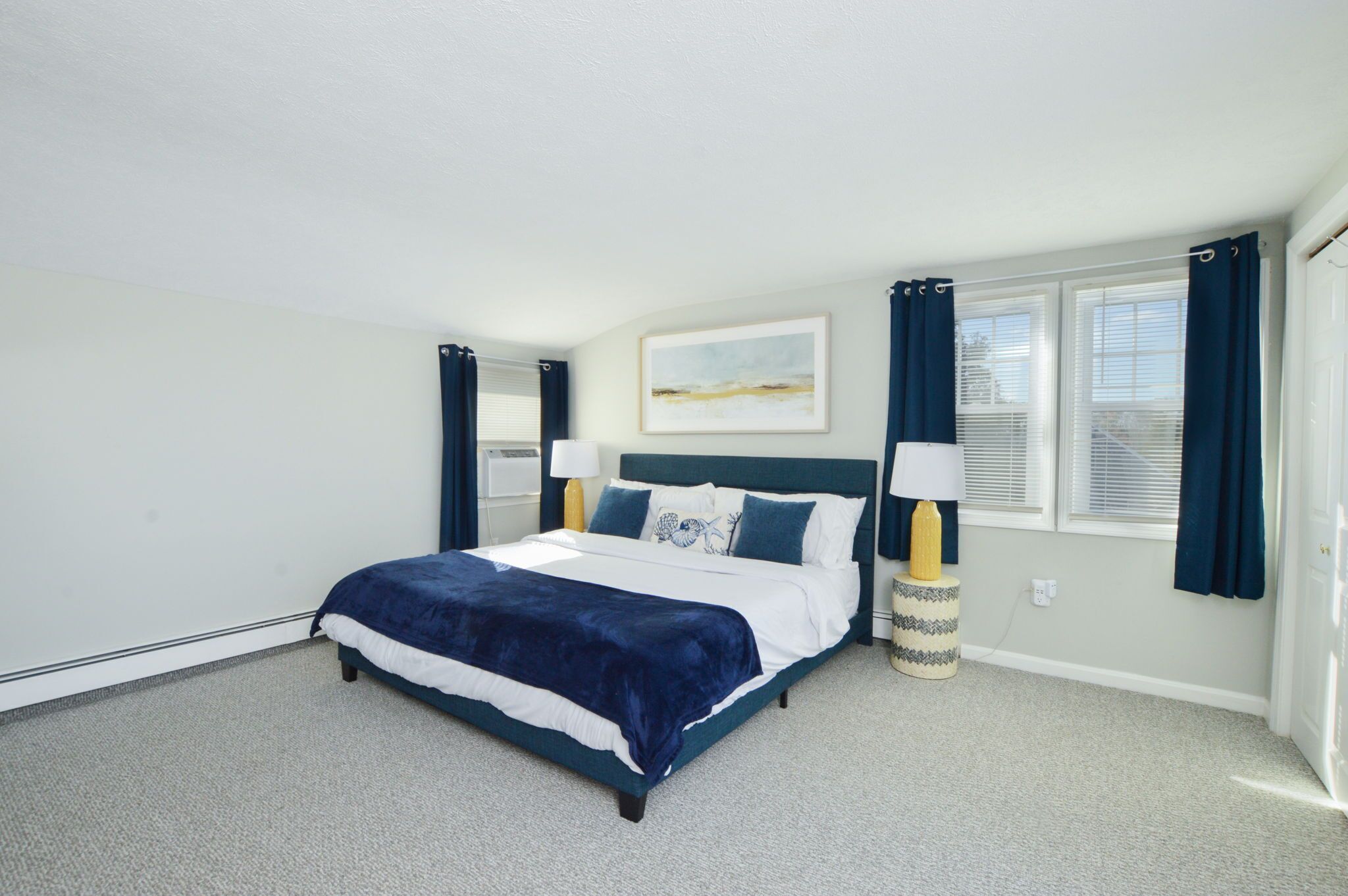 A bedroom with a bed, two bedside lamps, a window with curtains, and a painting on the wall.