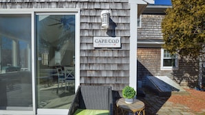 Une maison avec une porte-fenêtre coulissante, une chaise en osier avec un coussin vert et une petite table ronde sur laquelle est posée une plante en pot.