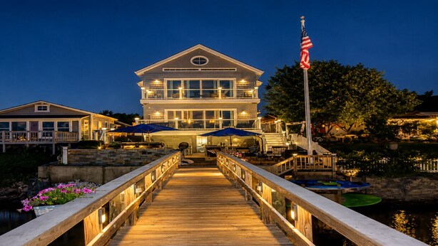 A waterfront property with a wooden deck leading to a large house, an American flag, and outdoor umbrellas. - Bella Vista, Luxury Waterfront Retreat Private Dock, Outdoor Living Near Newport (Portsmouth)