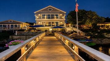 A waterfront property with a wooden deck leading to a large house, an American flag, and outdoor umbrellas.