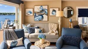 A living room with two blue armchairs, a beige sofa, and a round coffee table with a blue vase. There are framed pictures of seashells on the wall, and a view of the water is visible through the window. - Bella Vista, Luxury Waterfront Retreat Private Dock, Outdoor Living Near Newport (Portsmouth)