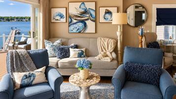 A living room with two blue armchairs, a beige sofa, and a round coffee table with a blue vase. There are framed pictures of seashells on the wall, and a view of the water is visible through the window.