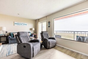 A room with a large window, two reclining chairs, a sofa, a small table, and a sign indicating a beach direction.