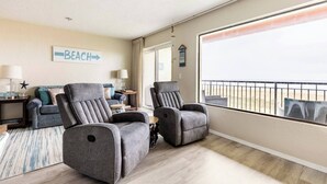 A spacious living room with two grey armchairs, a sofa, and a coffee table. A large window offers a view of the beach. - Sand Dollar Haven at Seaside Beach Club Unit 306 (Seaside)