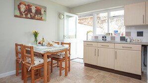 A kitchen with wooden cabinets, a dining table with chairs, a clock, and a window with a stone wall view. - 2 bedroom accommodation in Dawlish (Dawlish)