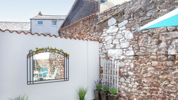 A courtyard with a stone wall, a white wall with a window, and potted plants. - 2 bedroom accommodation in Dawlish (Dawlish)