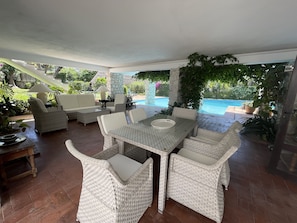 Ein überdachter Patio-Bereich mit Esstisch und Rattanmöbeln, mit Blick auf einen Swimmingpool.