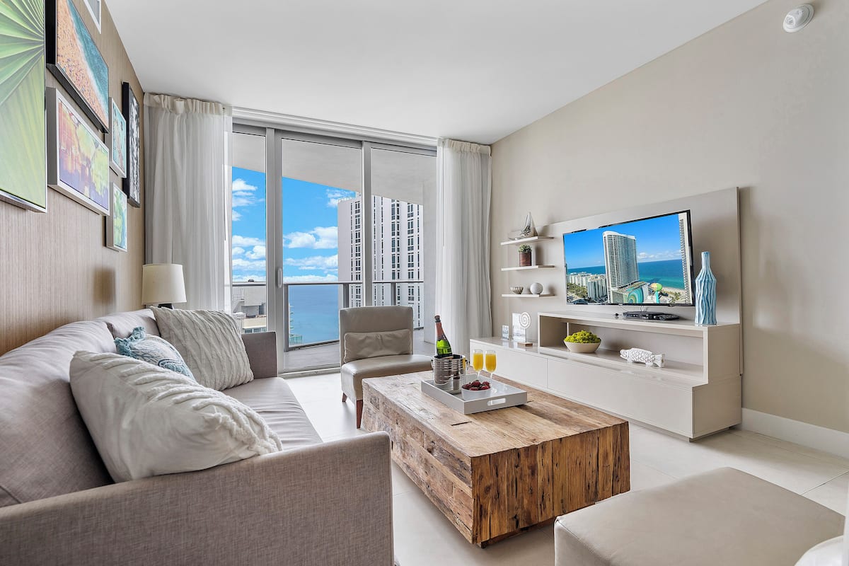A modern living room with a sofa, coffee table, and TV, featuring a cityscape view through large windows.