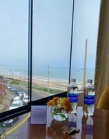 Quarto de hotel com vista para a praia e para a estrada. Há duas garrafas de água San Carlos sobre a mesa. Um vaso com flores também é visível.