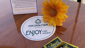 Pöydällä on Golden Mar -hotellin kyltti, kukka, hotellin logolla koristeltu alusta ja suklaapatukoita.