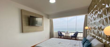 Habitación de hotel con una cama, televisor montado en la pared, vistas a la playa y una mesa con sillas.