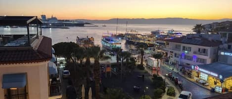 Un paisaje urbano costero al atardecer con un puerto deportivo, edificios y una calle con coches estacionados.