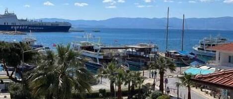 A coastal view with a large ship docked at the harbor, a marina with boats, and a parking area with cars.