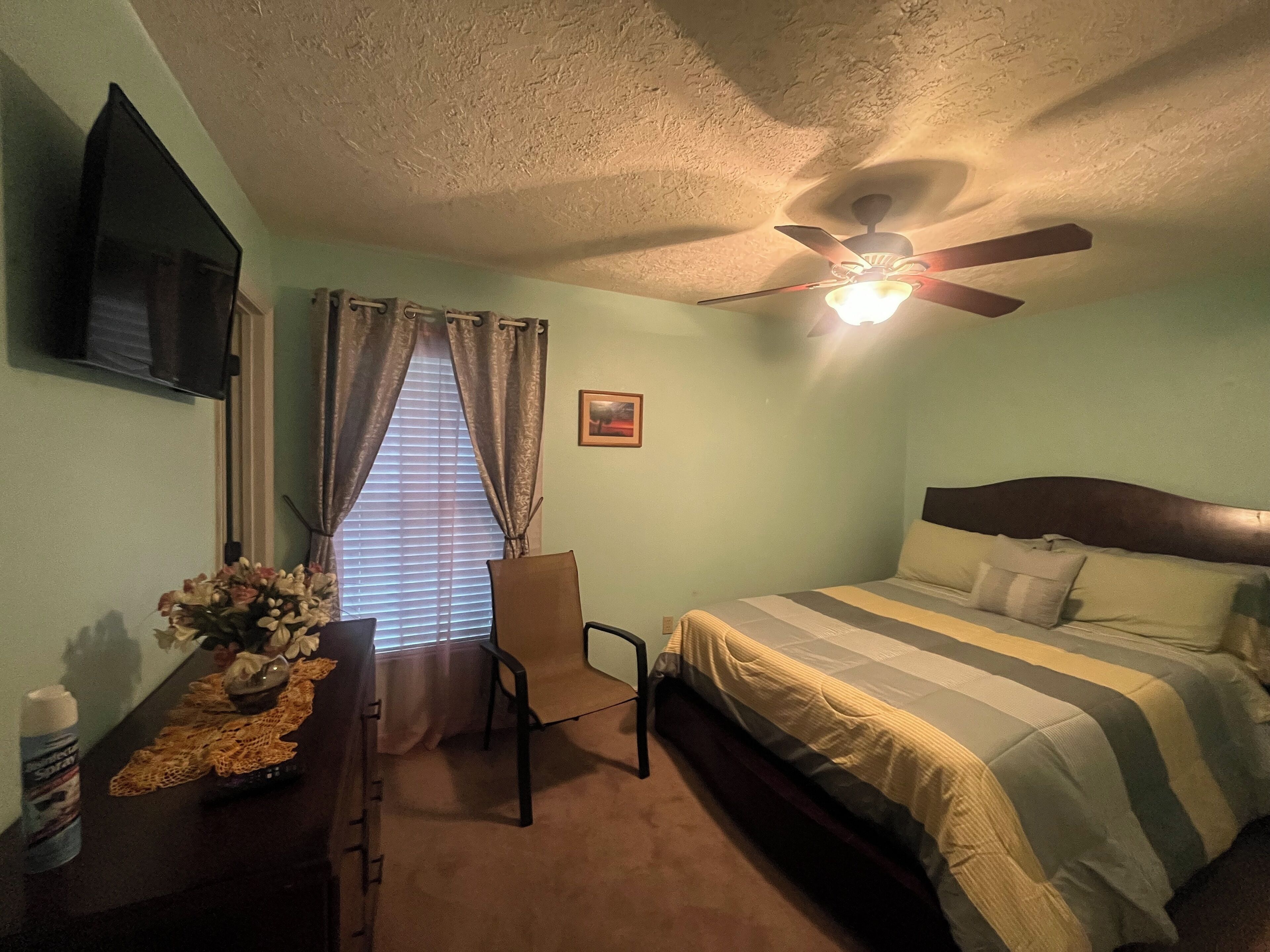 Une chambre à coucher comprenant un lit, un bureau, une chaise, un ventilateur de plafond, une fenêtre avec des rideaux et un téléviseur fixé au mur.