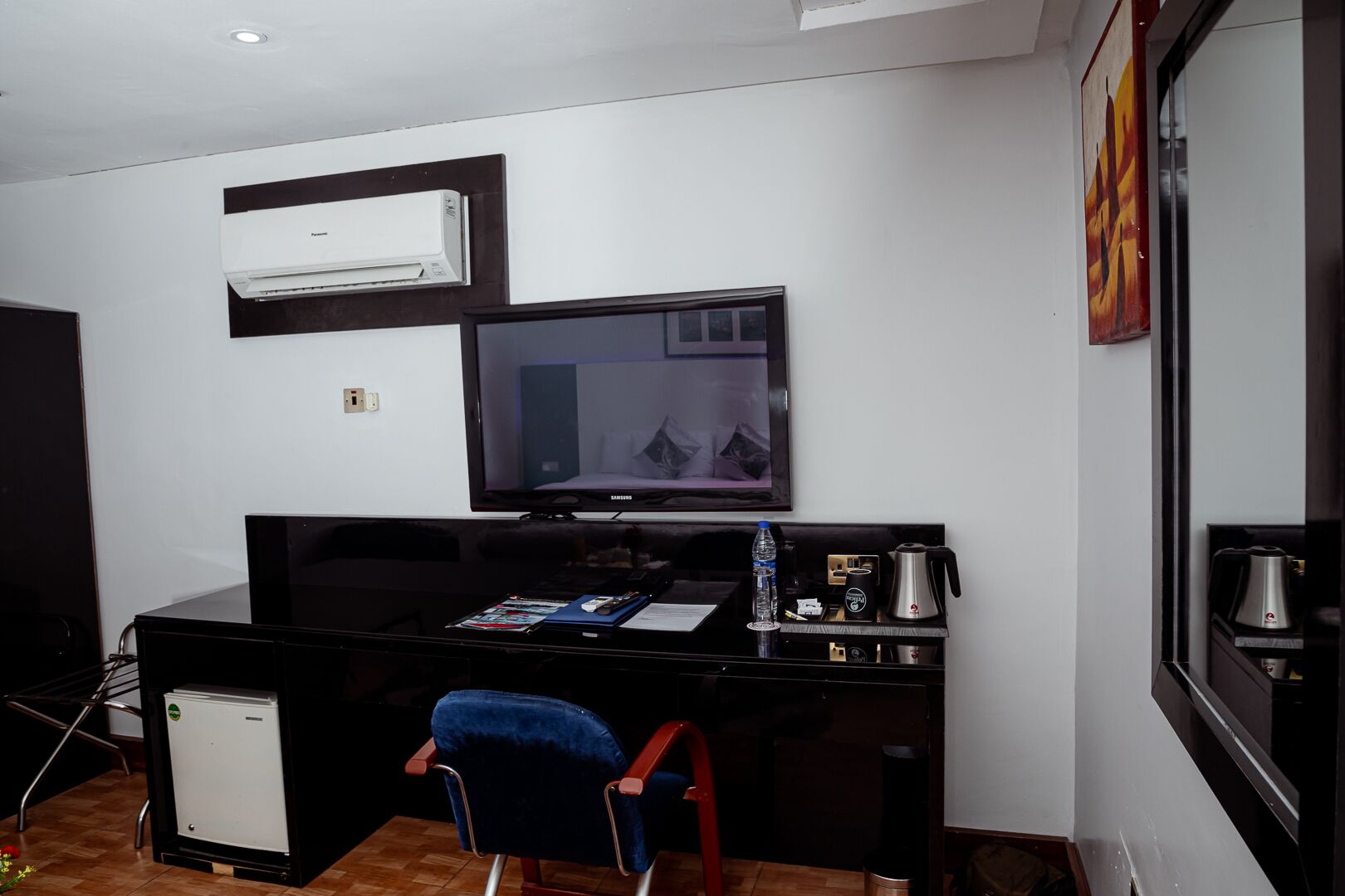 Une chambre d’hôtel de taille modeste, dotée d’un bureau noir, d’une télévision, d’un petit réfrigérateur et d’une chaise.