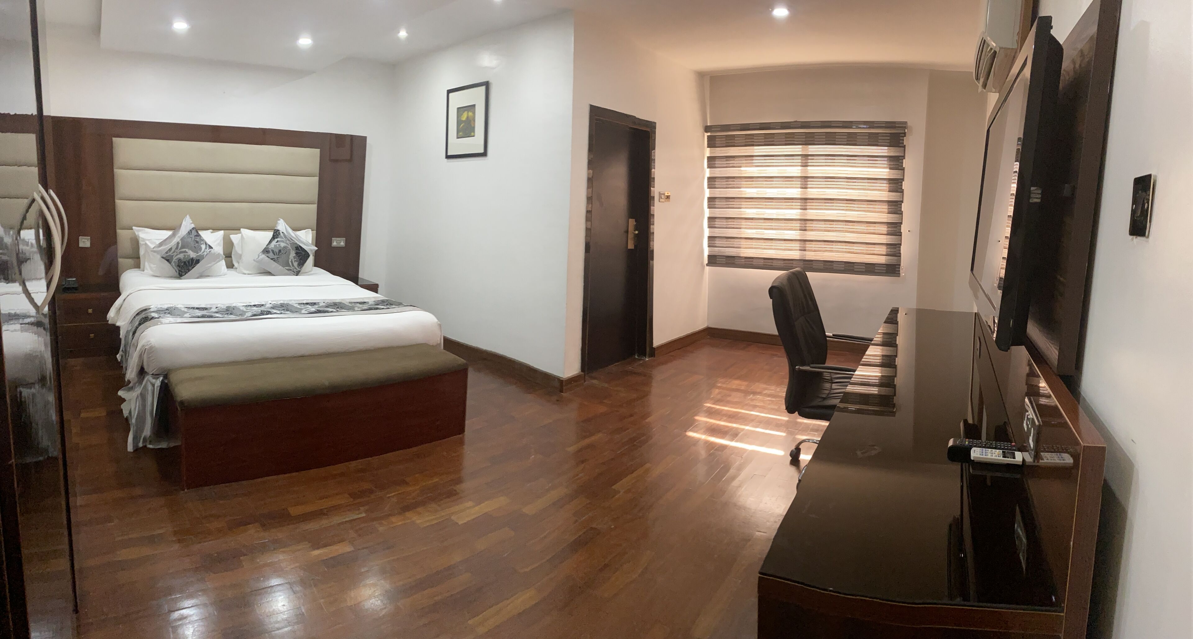 Ein Hotelzimmer mit einem Bett, einem Schreibtisch mit Flachbildfernseher, einem Sessel, einem Fenster mit Jalousien und einem Bild an der Wand.