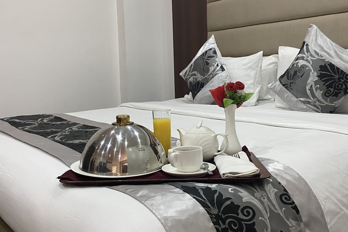 A neatly made bed with a tray of breakfast items, including a teapot, cup, and juice, and a decorative vase with a flower.