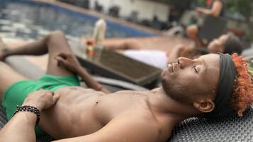 Una persona recostada en una silla de descanso junto a una piscina, con otra persona al fondo.