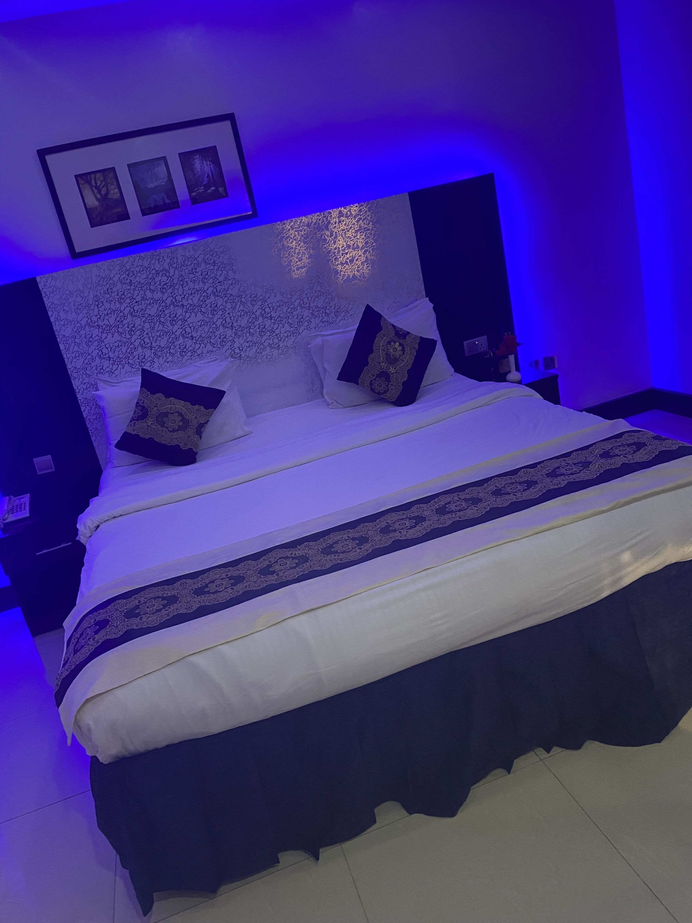 Cama bem arrumada com colcha branca e azul escura, acompanhada por duas mesas de cabeceira azuis escuras com abajures.