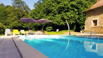 Une piscine entourée de verdure et d’un bâtiment en pierre, avec des chaises longues et des parasols à proximité.