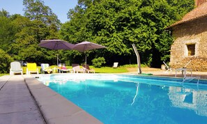 Une piscine entourée de verdure et d’un bâtiment en pierre, avec des chaises longues et des parasols à proximité.