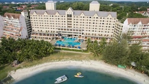 Un gran hotel con piscina central, rodeado de palmeras y una playa.