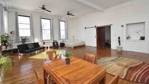Ein geräumiges Wohnzimmer mit einem Holzesstisch, einer Couch und einem Bett. Zwei Deckenventilatoren, große Fenster und eine Schiebetür zu einem anderen Raum sind vorhanden.
