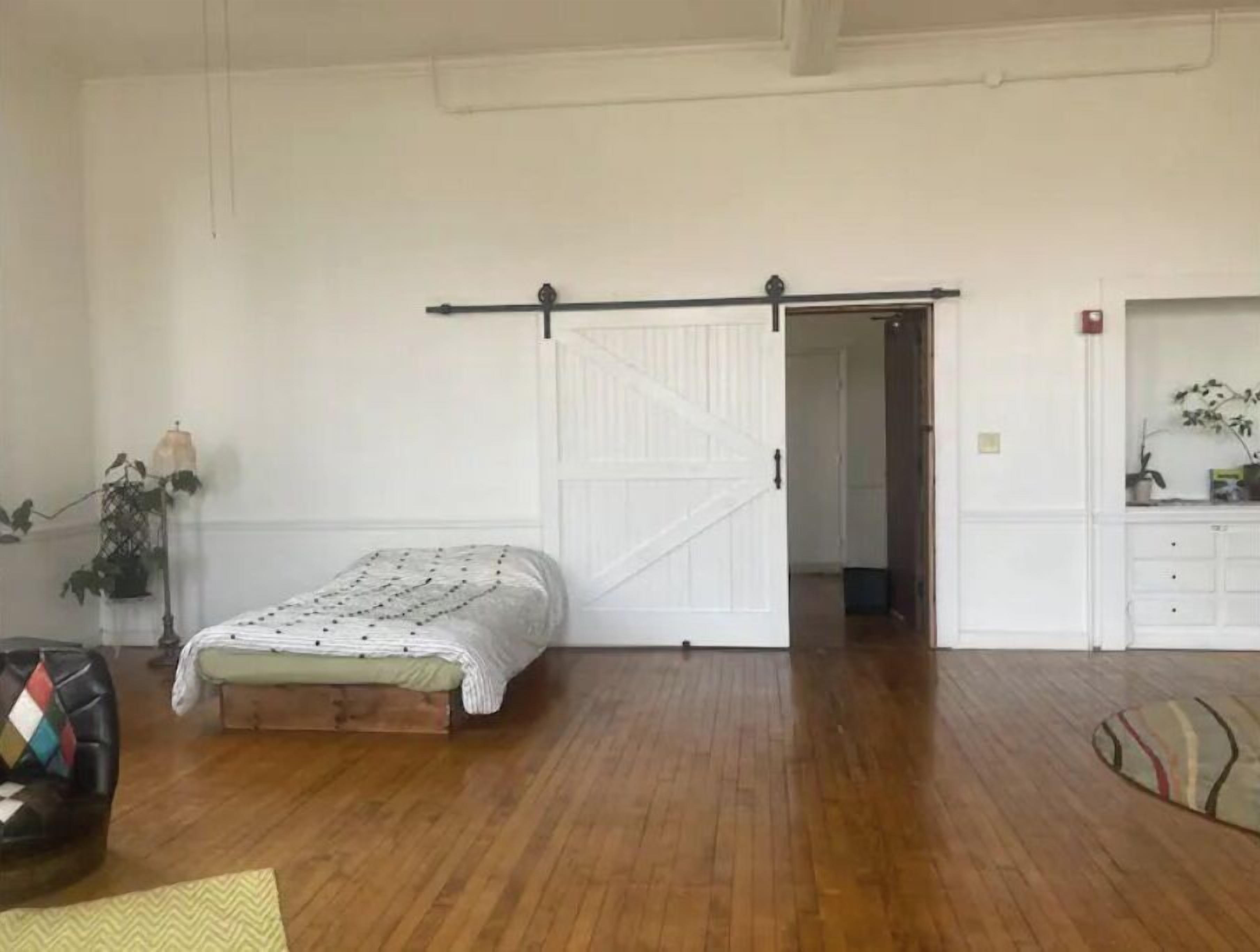 A spacious room with a wooden floor, a bed with a patterned bedspread, a sliding door, a small desk with a chair, and a potted plant.