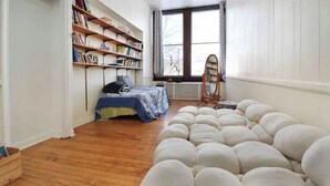 A bedroom with a wooden floor, a bed with a blue cover, a bookshelf, a window with curtains, and a white pouf. - Creative living 4 (Youngstown)