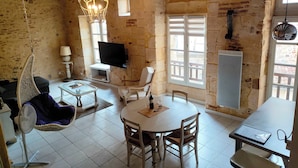 A rustic room with exposed brick walls, a hanging chair, a dining table with chairs, a TV mounted on the wall, and a window with shutters.