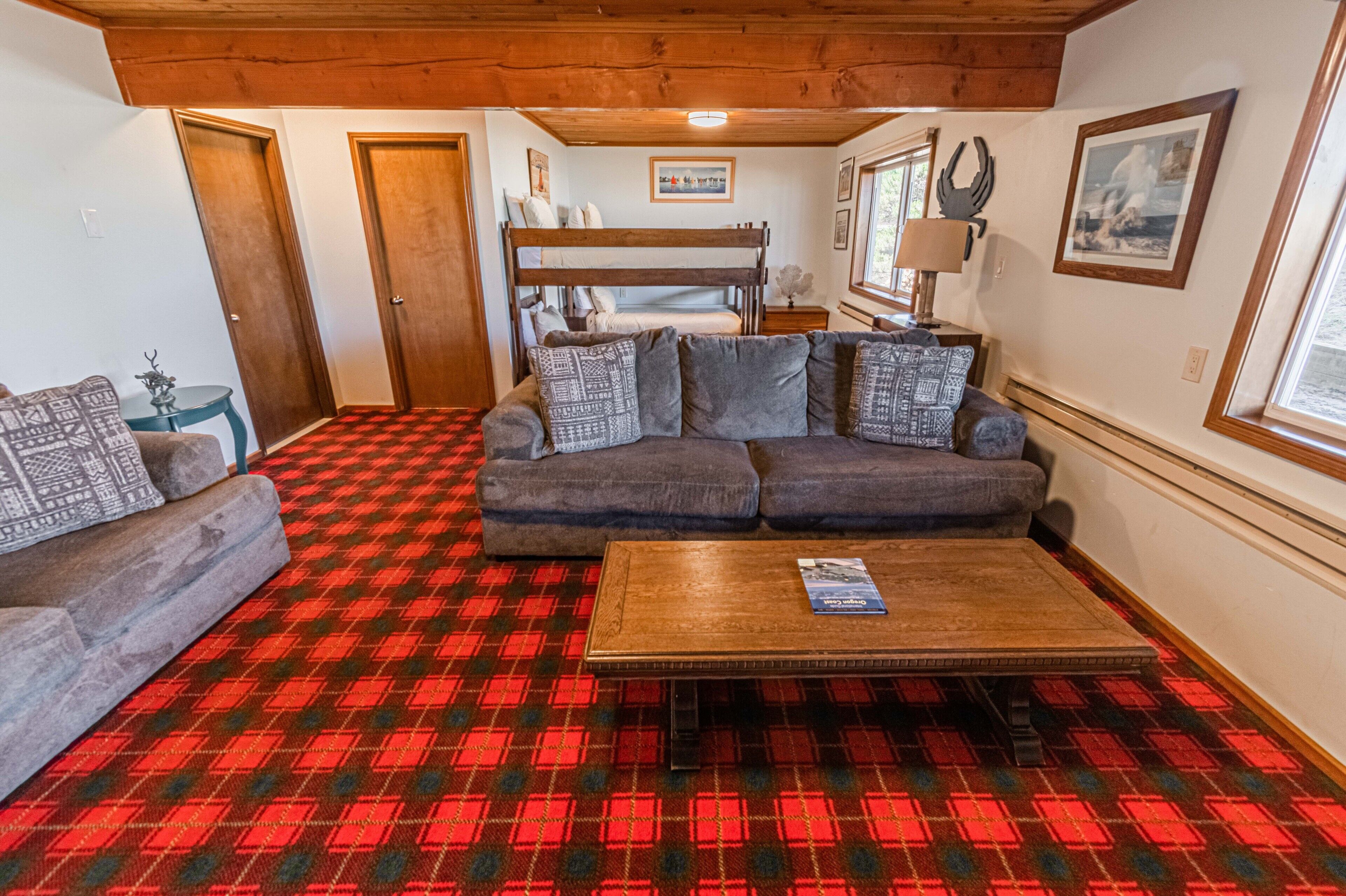 A cozy living room with a checkered carpet, a sofa set, a coffee table, and a bunk bed in the background.