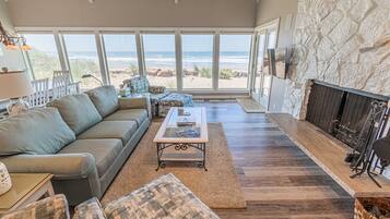 A spacious living room with a large window overlooking the beach, a stone fireplace, and comfortable seating.
