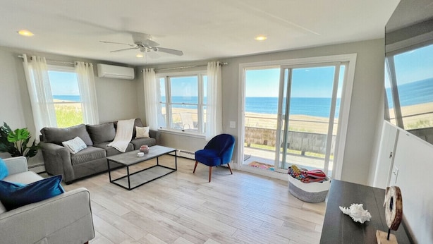 Een ruime woonkamer met een grijze bank, een blauwe fauteuil en een salontafel. Grote ramen bieden uitzicht op het strand en de oceaan.