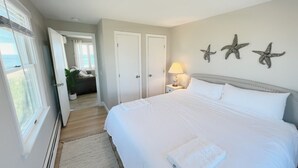 Een slaapkamer met een groot bed, een nachtkastje met lamp, decoratie van zeesterren en uitzicht op de zee door een geopende deur.