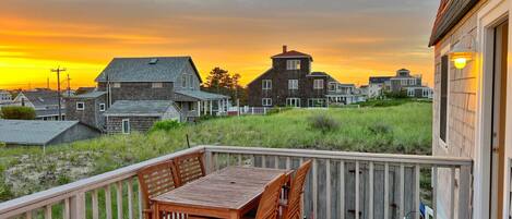 A wooden deck with a table and chairs, overlooking a grassy area and buildings during sunset.