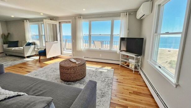 Un salon spacieux avec un grand canapé gris, un pouf en osier et un téléviseur à écran plat posé sur un meuble blanc. La pièce est dotée de grandes fenêtres offrant une vue sur la plage et l’océan.