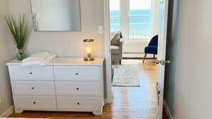 Une chambre avec un grand lit, une commode à tiroirs, une lampe de chevet et une vue sur le balcon et l’océan, visible par la porte ouverte.