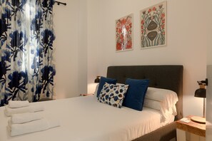 Una cama bien hecha con sábanas blancas, almohadas azules y estampadas, y un cabecero oscuro.