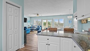 A spacious kitchen with white cabinets, a granite countertop, and a view of the living area with a blue sofa and wicker furniture. - Duplex with private pool and easy beach access,in the Spinnaker's Reach (Emerald Isle)