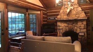 Interior acogedor de una cabaña con chimenea de piedra, vigas de madera, un televisor y vistas por la ventana.