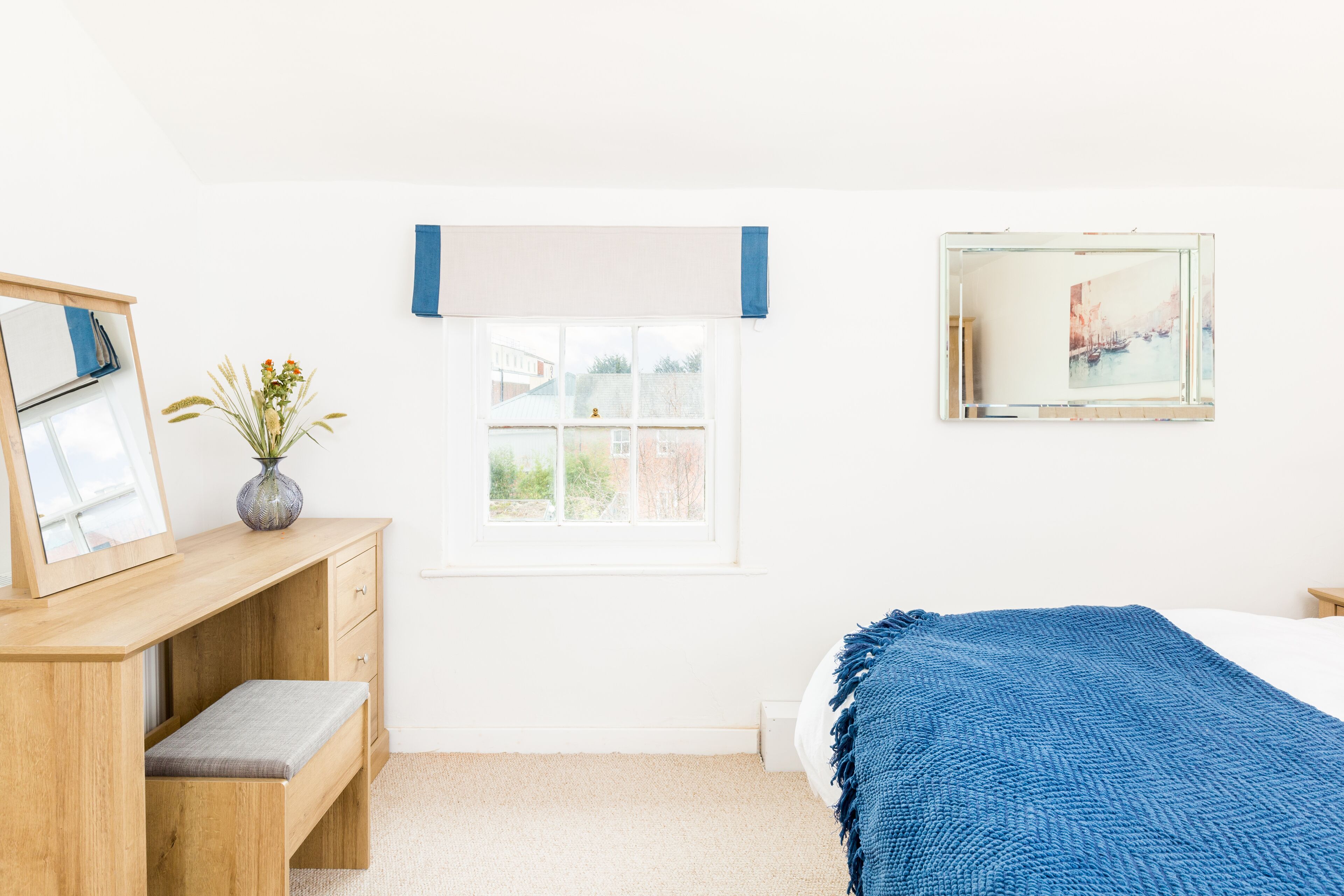 Une chambre avec un bureau en bois, une fenêtre avec des stores, un lit recouvert d’une couverture bleue et un tableau accroché au mur.